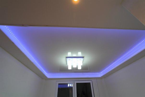 Podwieszany sufit z oświetleniem LED w kolorze niebieskim, centralnie umieszczona nowoczesna lampa sufitowa z białymi kloszami, widoczne okno w tle.