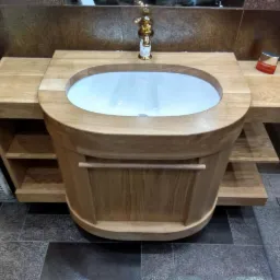 Owalna umywalka ceramiczna w drewnianej obudowie z półkami, złota bateria, brązowe płytki na ścianie i podłodze.