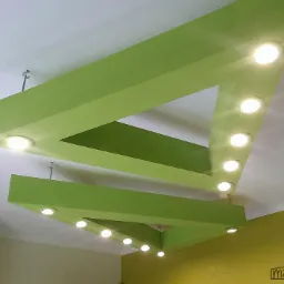 Awangardowy sufit podwieszany z płyty kartonowo-gipsowej w kształcie dwóch trójkątów, pomalowany na jasnozielono, z wbudowanymi okrągłymi lampami LED.