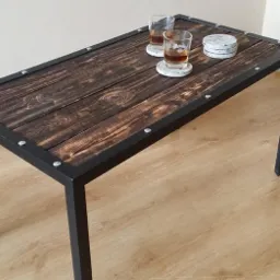 Stolik kawowy z blatem z ciemnego, postarzanego drewna i metalową ramą, na blacie dwa drinki w szklankach i podkładki, widoczne logo firmy w rogu.