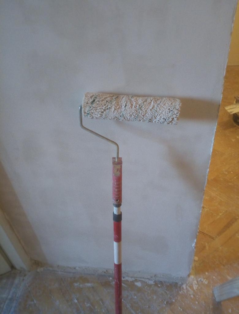 Świeżo pomalowana ściana z wałkiem do malowania na kiju, widoczne niedoskonałości na powierzchni ściany i ślady farby na podłodze.