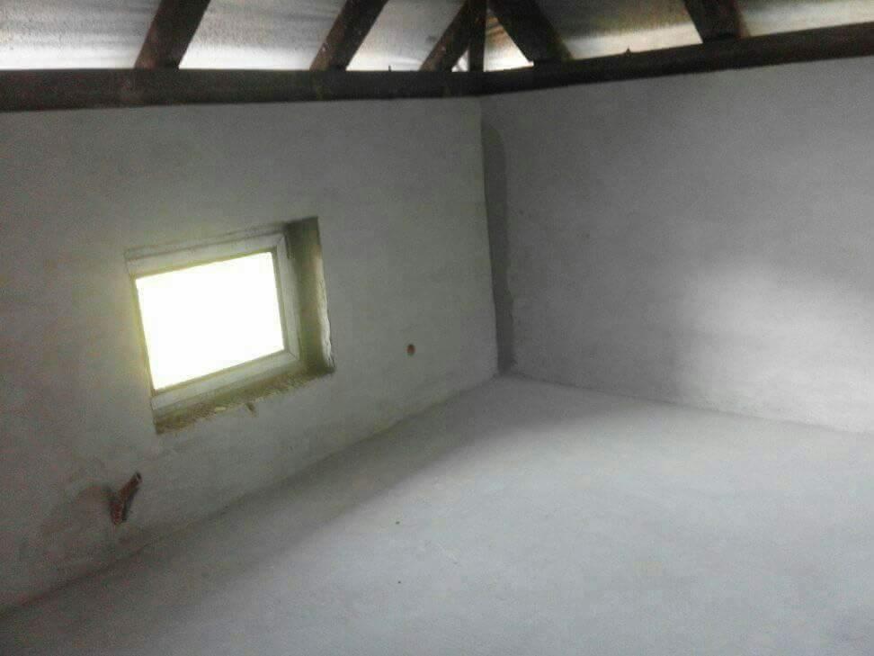 Poddasze po wylewce posadzki i wstępnym otynkowaniu ścian, widoczne drewniane elementy konstrukcji dachu i okno z widocznym światłem dziennym.