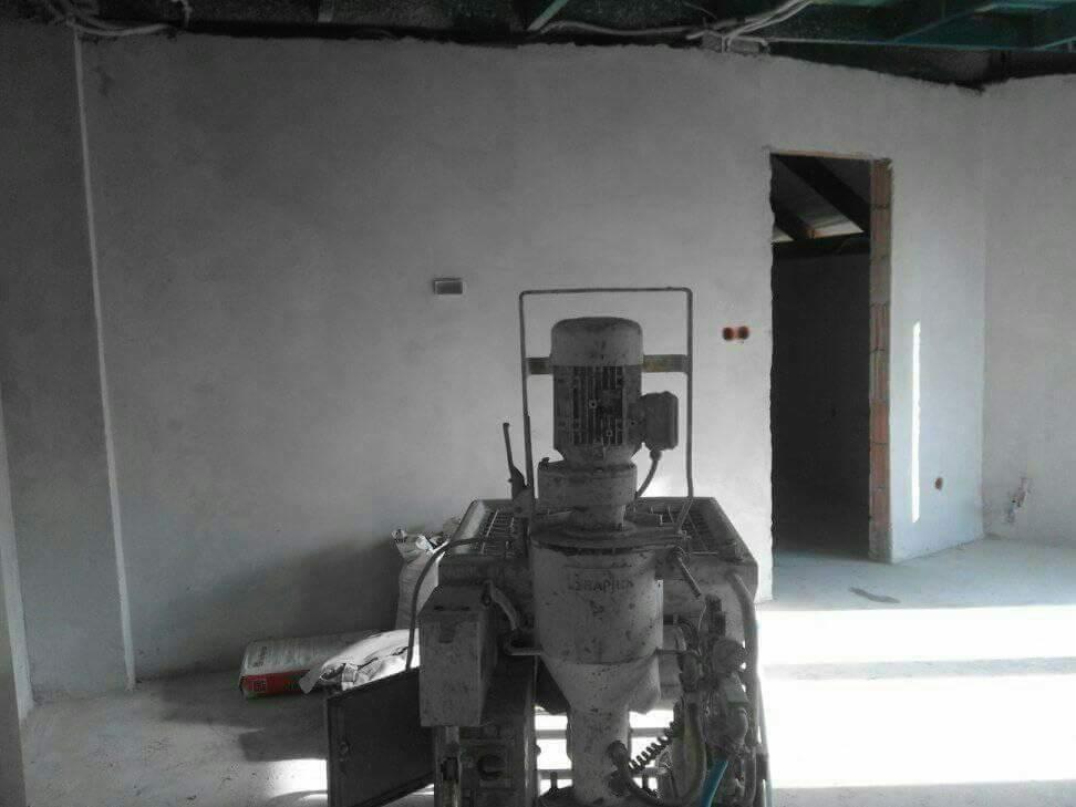 Wnętrze w trakcie tynkowania maszynowego; widoczna maszyna do tynkowania na tle świeżo otynkowanej ściany i surowego otworu drzwiowego.