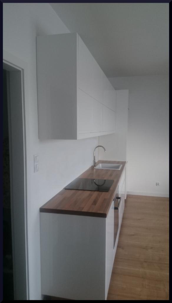 Nowoczesna kuchnia z białymi szafkami wiszącymi i blatem z drewna, widok z boku, z widoczną płytą indukcyjną i zlewem ze stali nierdzewnej.