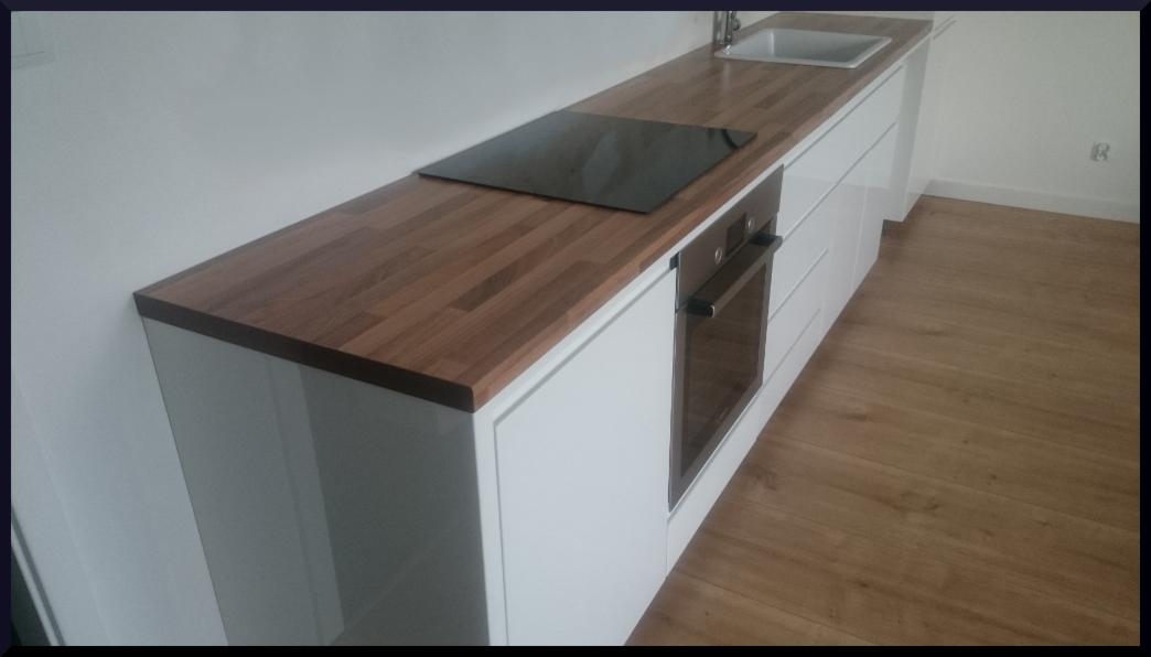 Nowoczesna kuchnia z białymi szafkami i blatem z drewna w odcieniach brązu, na którym umieszczona jest czarna płyta indukcyjna i zlewozmywak, a pod nim piekarnik.