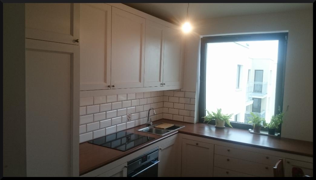 Biała kuchnia z górnymi szafkami, białymi płytkami metra i drewnianym blatem, okno z roślinami w doniczkach, widok na budynek z balkonami.