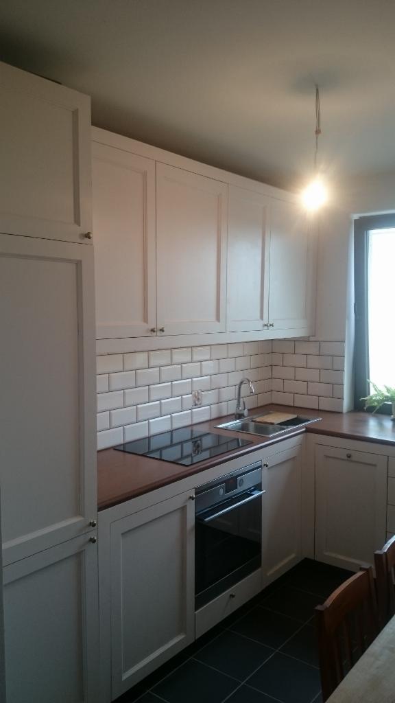 Jasna kuchnia z białymi szafkami, brązowym blatem i białymi płytkami na ścianie, widoczny zlew, płyta indukcyjna i piekarnik