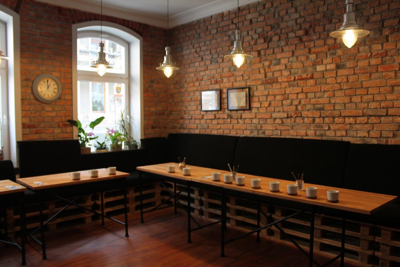 Jasne wnętrze kawiarni z ceglaną ścianą, drewnianymi stołami na bazie palet i czarnymi kanapami wzdłuż ściany, z filiżankami i akcesoriami na stołach.