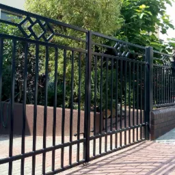 Nowoczesna, czarna brama dwuskrzydłowa z metalowymi prętami i dekoracyjnymi rombami w górnej części, prowadząca do posesji z bujną roślinnością, widoczna skrzynka pocztowa z numerem 35.