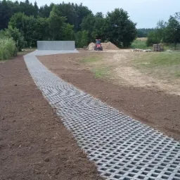 Długa ścieżka z szarej ażurowej kostki prowadzi przez teren budowy, w tle widoczny mur z betonowych bloków, traktor i materiały budowlane.