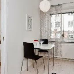 Jasne, minimalistyczne wnętrze: otwarta przestrzeń z aneksem kuchennym, stołem jadalnym i widokiem na sypialnię z łóżkiem ozdobionym czerwonymi poduszkami i artystycznym obrazem kwiatowym.