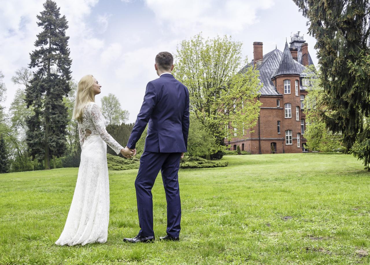 Para młoda trzymająca się za ręce na tle rozległego trawnika i ceglanego dworku z wieżyczkami w stylu neogotyckim.