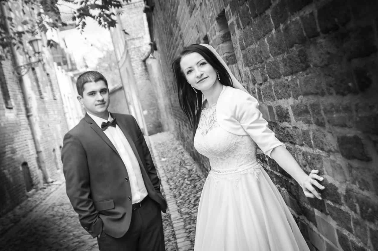 Czarno-białe zdjęcie pary młodej w wąskiej uliczce z ceglanymi ścianami. Panna młoda w koronkowej sukni i bolerku, z welonem i kolczykami, opiera dłoń o mur. Pan młody w garniturze z muchą stoi...