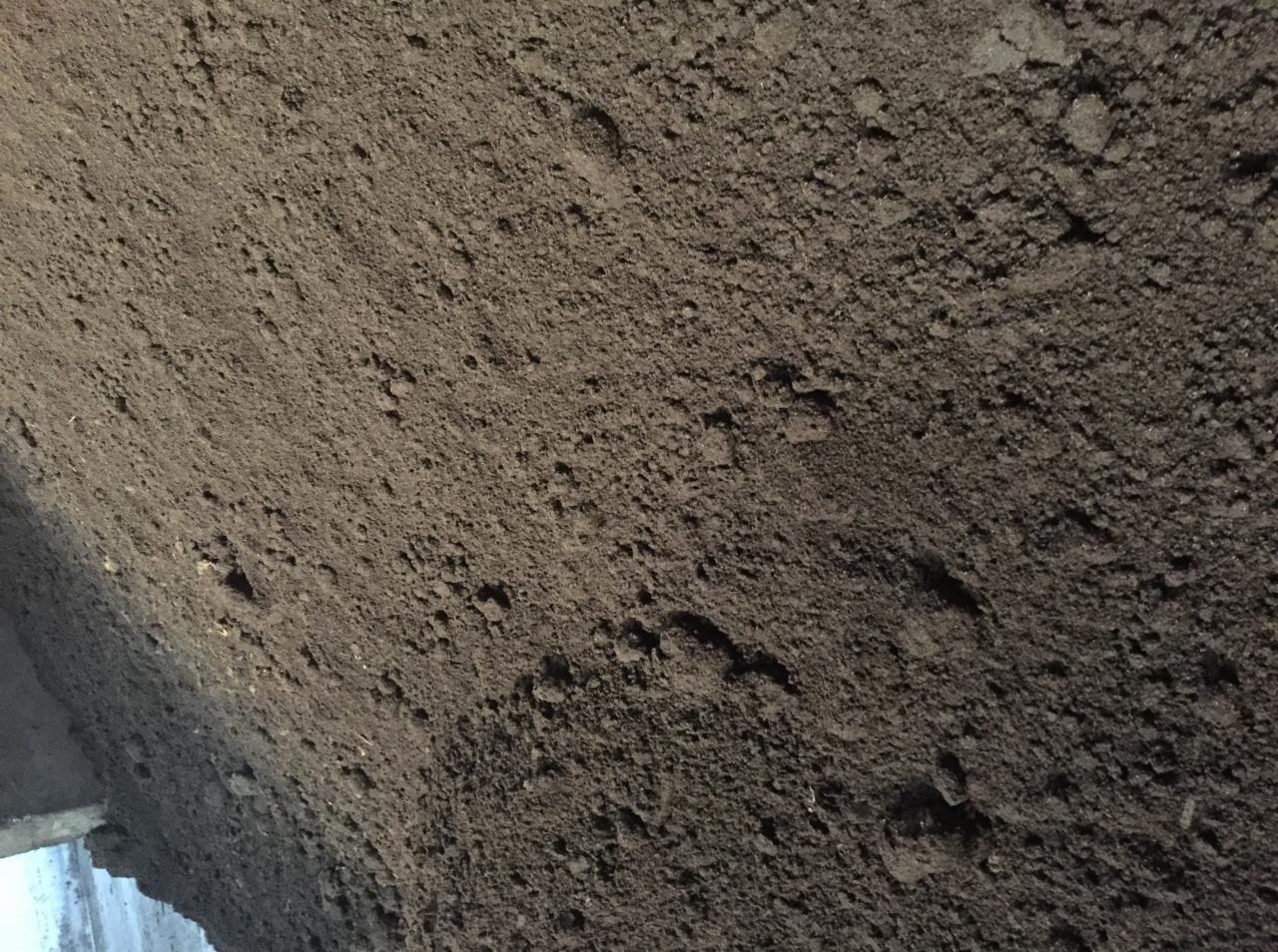 Sterta ciemnej, sypkiej ziemi z widocznymi grudkami, oświetlona z góry, z fragmentem betonowej konstrukcji w lewym dolnym rogu.