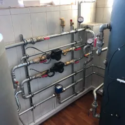 Rozbudowana instalacja hydrauliczna w kotłowni, widoczne liczne rury, zawory, manometry, pompy obiegowe i zasobnik ciepłej wody, zamontowane na ścianie z białych płytek.