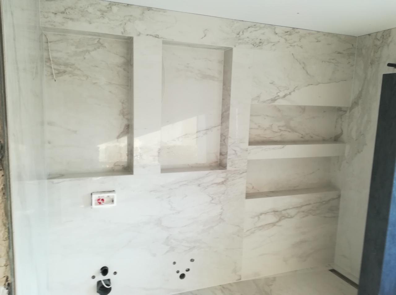 Ściana łazienki wykończona płytami imitującymi marmur, z wnękami pełniącymi funkcję półek, widoczne instalacje hydrauliczne przed wykończeniem.