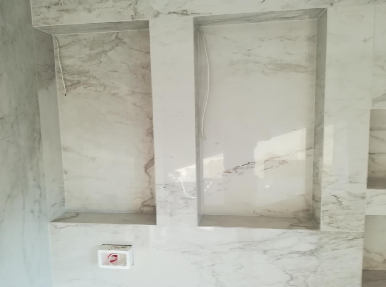 Ściana łazienki wyłożona marmurowymi płytami z wnękami, widoczne przewody elektryczne w jednej z wnęk oraz gniazdko elektryczne.