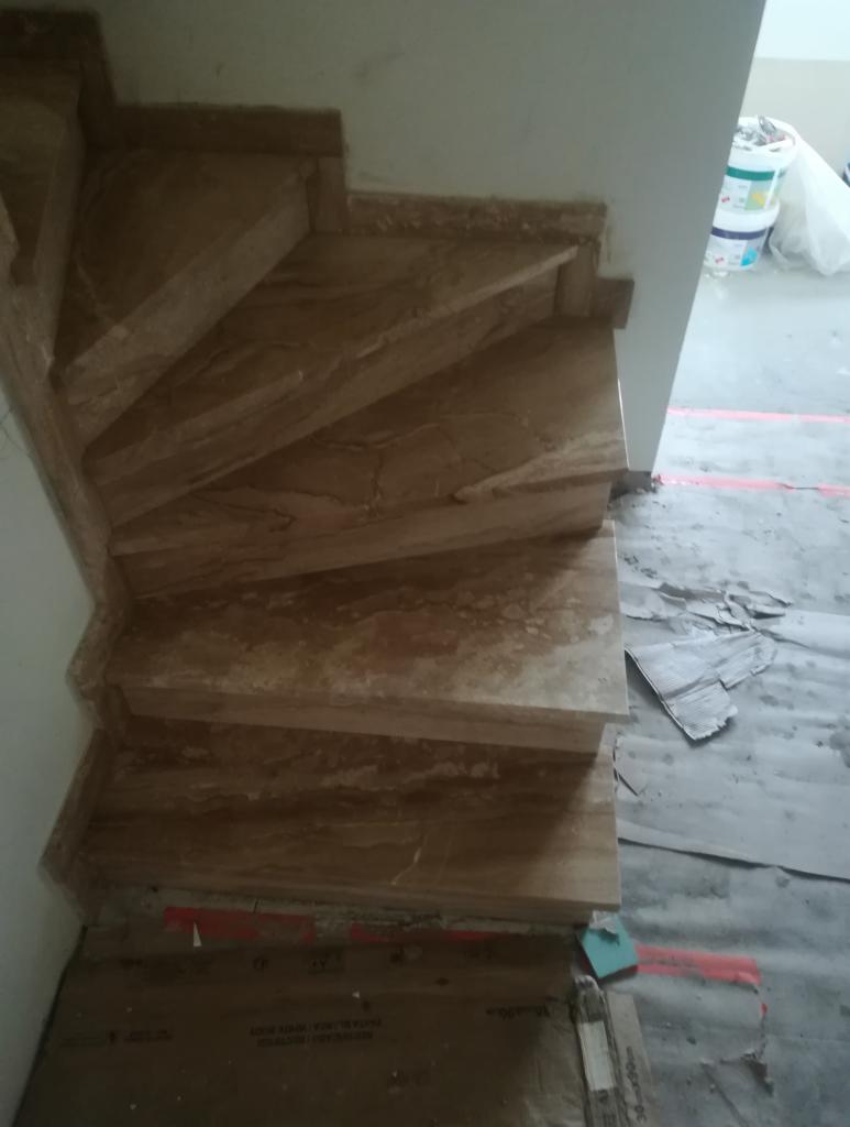 Kątowe schody kamienne z beżowego trawertynu, widoczne podczas montażu, z zabezpieczonym podłożem kartonem i folią.