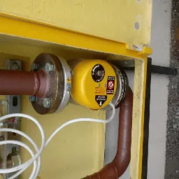 Żółta skrzynka gazowa z widoczną instalacją, zaworem z napisem 'Zamykanie Ręczne' i rurami w kolorze miedzianym, zamontowana na zewnątrz budynku.