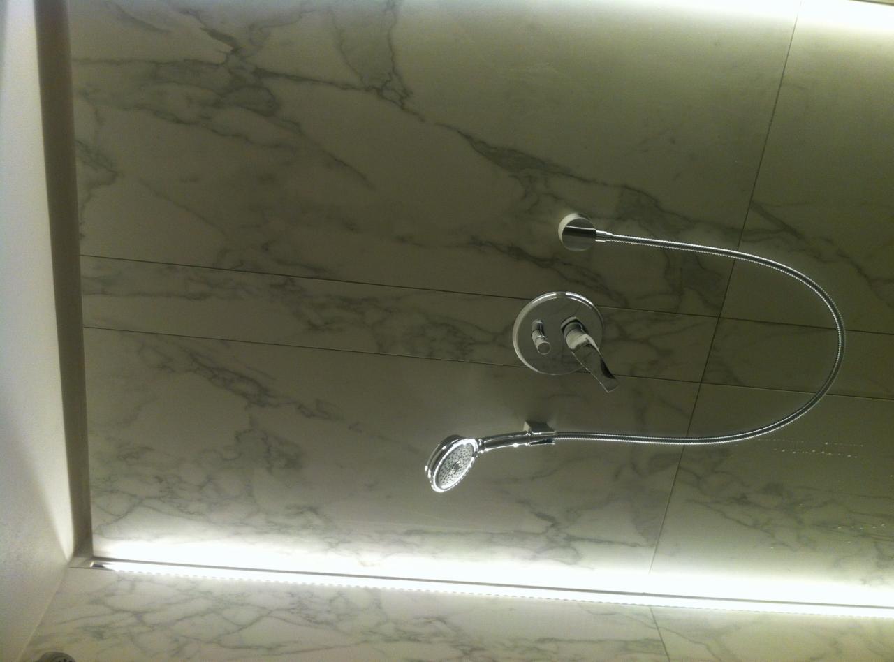 Biała marmurowa ściana kabiny prysznicowej z chromowaną baterią i słuchawką prysznicową na elastycznym wężu, oświetlona listwą LED u dołu kadru.