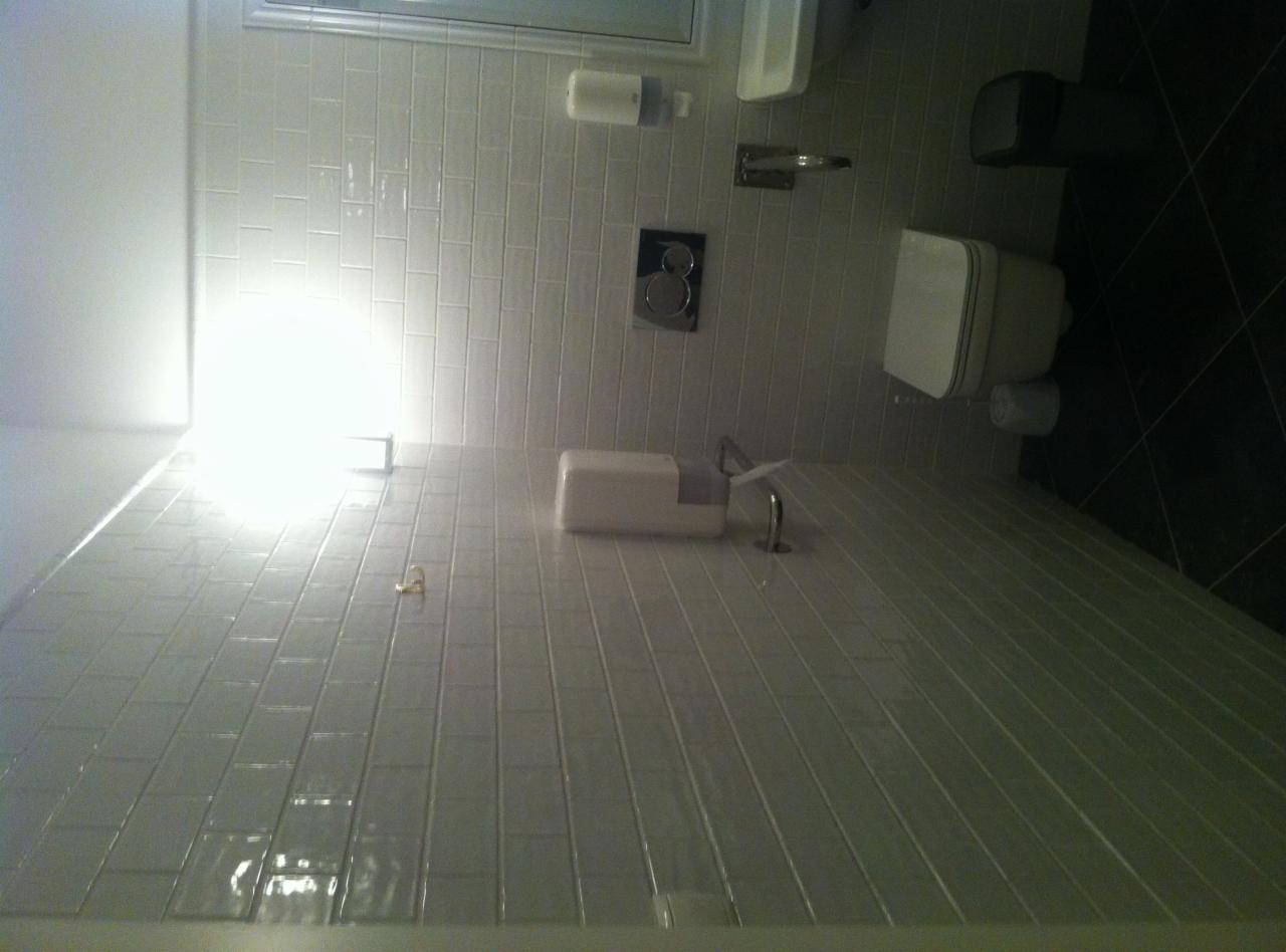 Jasne, wykafelkowane wnętrze łazienki z umywalką, toaletą i dozownikiem mydła, sfotografowane pod kątem, ukazujące czarne płytki podłogowe.