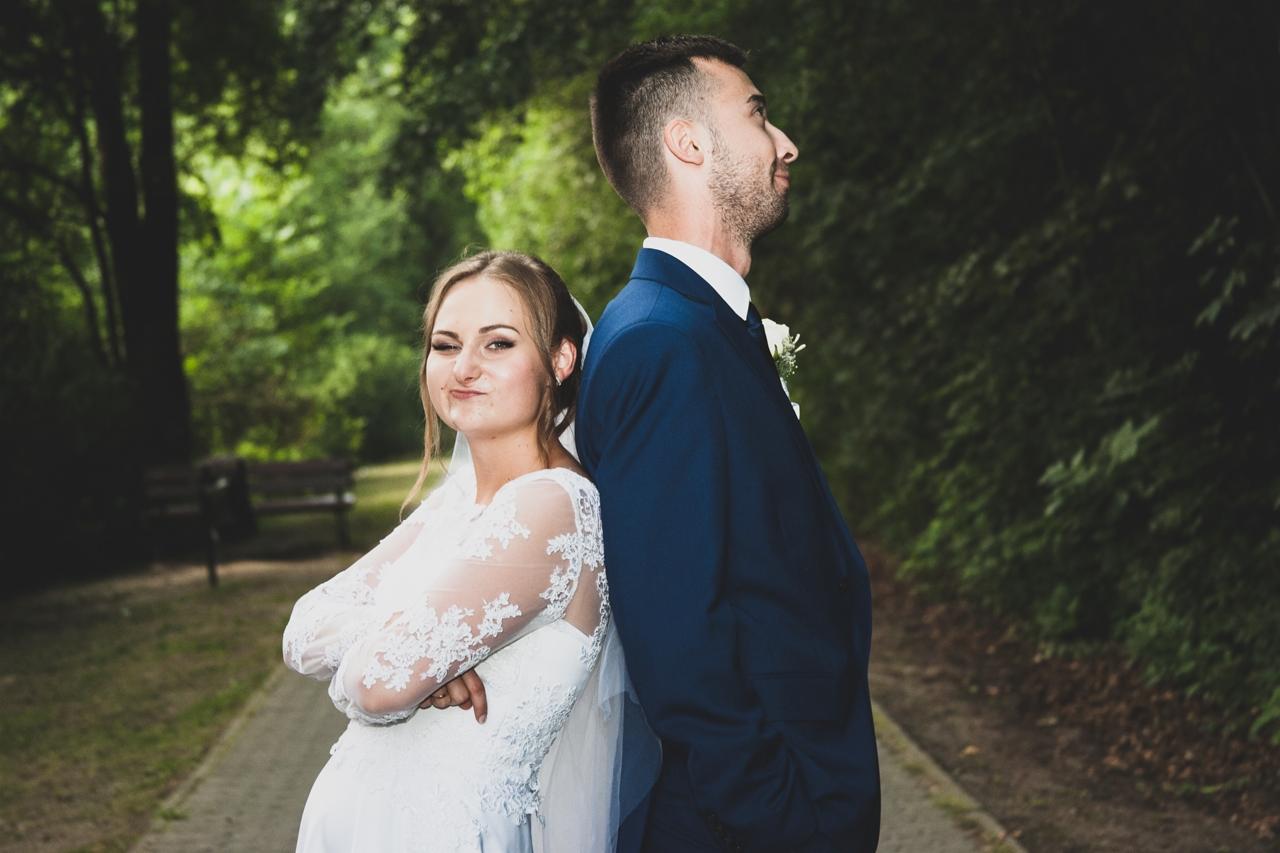 Para młoda stoi plecami do siebie na ścieżce w parku: panna młoda z przymrużonym okiem, pan młody patrzy w górę.