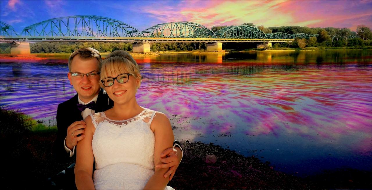 Para młoda, on w czarnym garniturze z fioletową muchą, ona w białej sukni ślubnej z koronką, pozują na tle mostu i rzeki z kolorowymi refleksami.