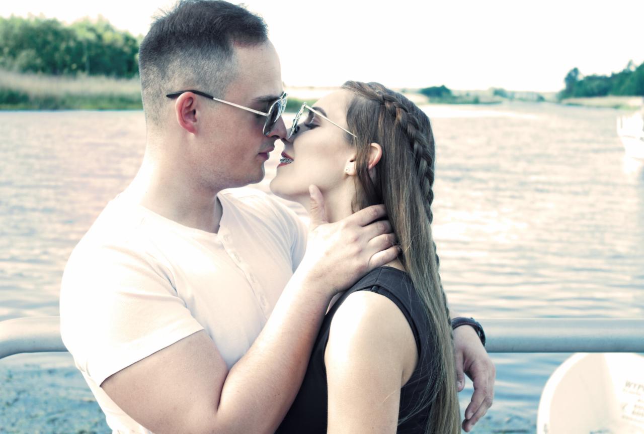 Młoda para w okularach przeciwsłonecznych, w zbliżeniu, prawie się całuje na tle wody i zieleni.