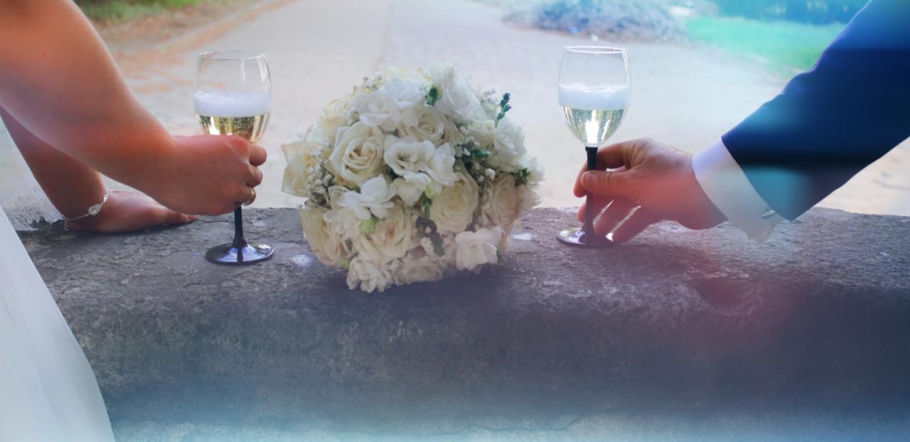 Para wznosi toast szampanem, bukiet z białych róż leży na kamiennym murku, widoczne fragmenty sukni panny młodej i garnituru pana młodego.