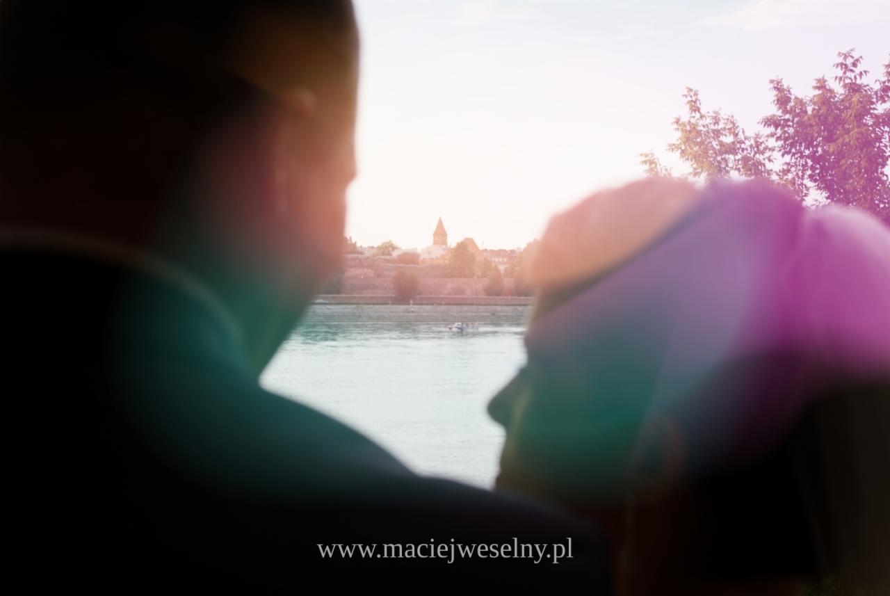 Para młoda na tle rzeki i miasta z wieżą kościoła, ujęcie zza ramion, delikatne rozmycie, logo fotografa na dole.