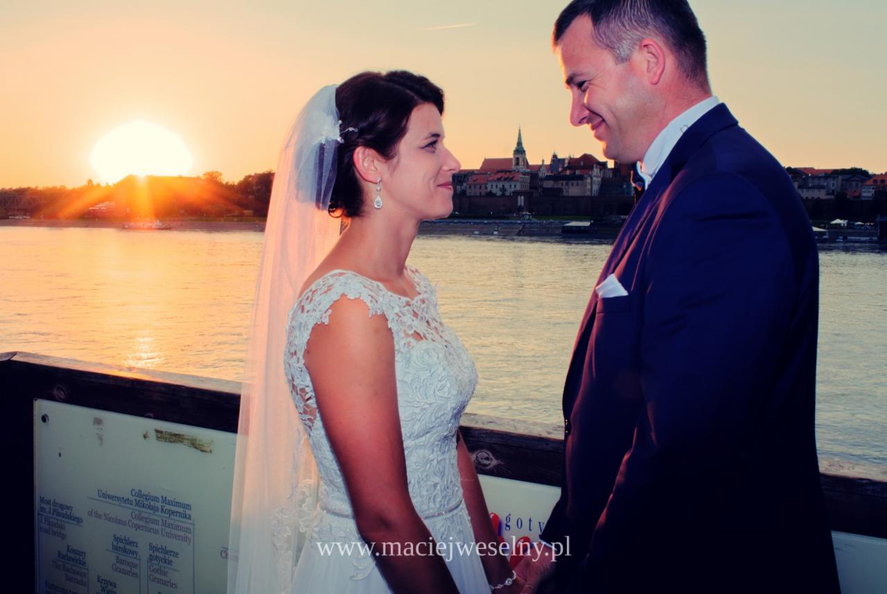 Para młoda, panna młoda w koronkowej sukni i welonie, pan młody w granatowym garniturze, patrzą na siebie na tle zachodzącego słońca nad rzeką i panoramą miasta z wieżą kościelną.