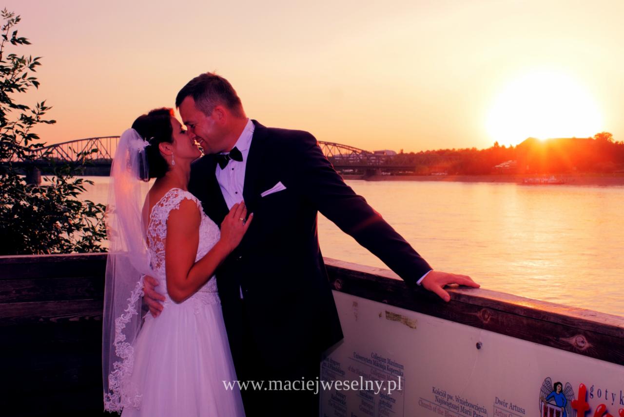 Pocałunek nowożeńców na tle zachodzącego słońca nad rzeką, w tle widoczny most i tablica informacyjna z elementami gotyckimi.