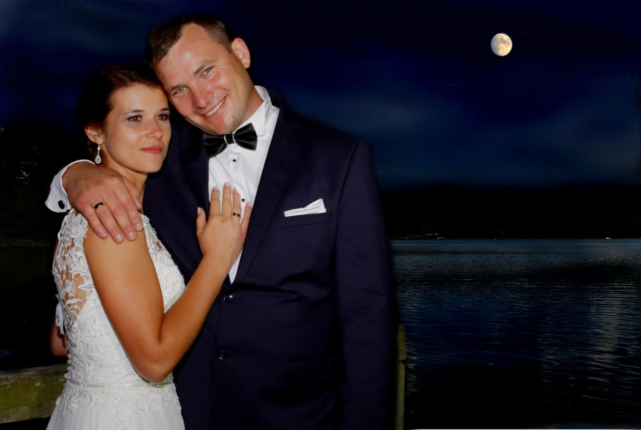 Para młoda w eleganckich strojach, obejmująca się na tle nocnego jeziora i księżyca w pełni, pierścionki na palcach wskazują na zawarcie małżeństwa.