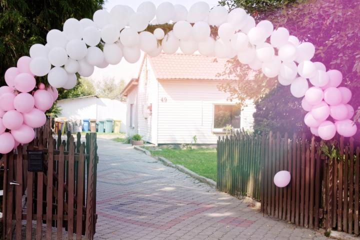 Brama wejściowa udekorowana łukiem z białych i różowych balonów, w tle biały dom z czerwonym dachem.