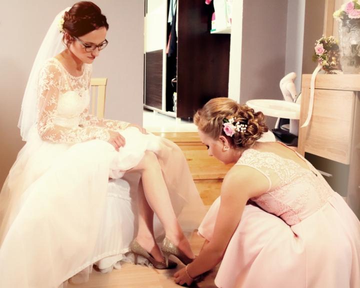Panna młoda w okularach, w koronkowej sukni ślubnej, siedzi podczas gdy druhna zapina jej błyszczące, złote szpilki.