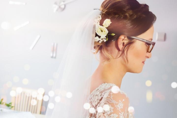 Panna młoda w okularach, z welonem i kwiatami we włosach, profil, delikatny makijaż, koronkowy dekolt sukni ślubnej.