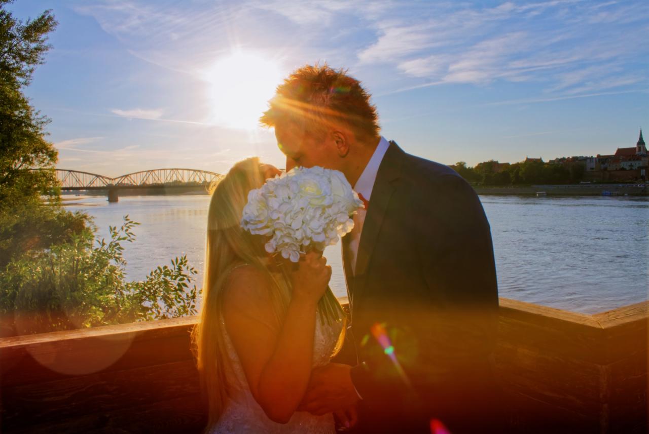 Para młoda całuje się na tle rzeki i mostu, oświetlona promieniami zachodzącego słońca, panna młoda trzyma bukiet białych kwiatów.