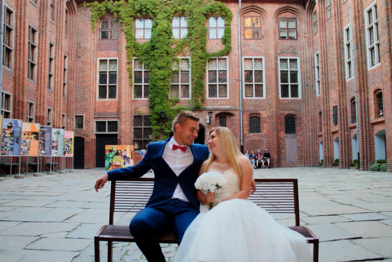 Uśmiechnięta para młoda siedzi na ławce na tle ceglanego dziedzińca z bluszczem, panna młoda trzyma bukiet z białych kwiatów, pan młody w granatowym garniturze i czerwonej muszce.