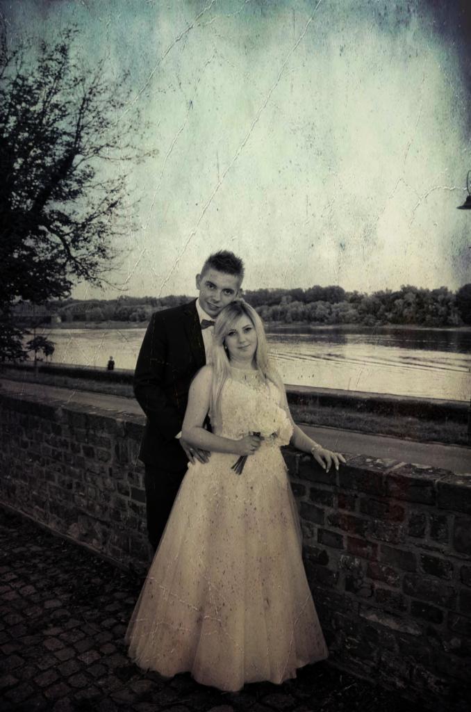 Para młoda w stylizowanej sesji plenerowej: on w czarnym garniturze z muchą, ona w jasnej sukni ślubnej z bukietem, pozują na tle ceglanego murku z rzeką w tle, efekt postarzanej fotografii.