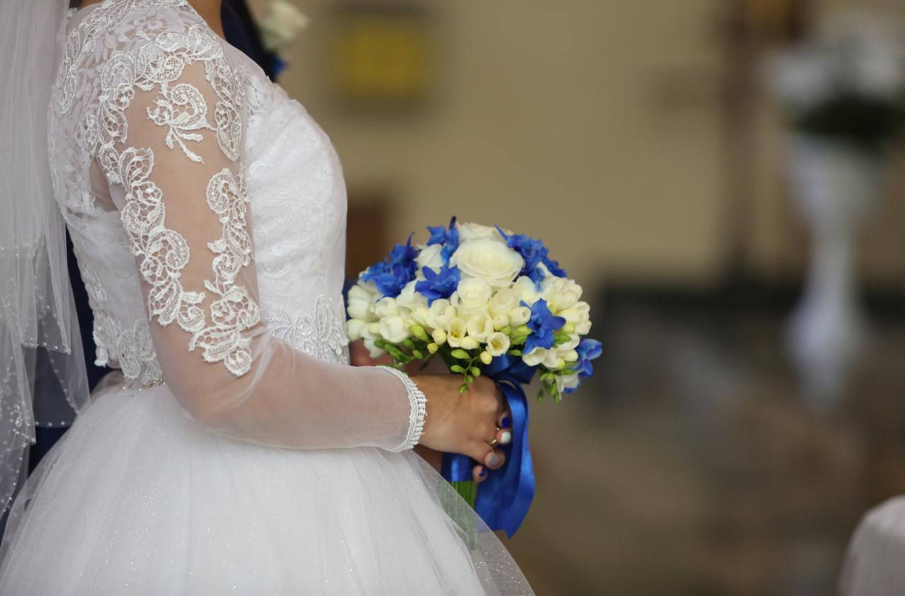 Panna młoda w koronkowej sukni trzyma bukiet białych i niebieskich kwiatów, zapięty niebieską wstążką, bransoletka na nadgarstku.