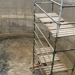 Wnętrze betonowego zbiornika z metalowym rusztowaniem, widoczne ślady wilgoci i zacieków na ścianach i podłożu, na zewnątrz zielony krajobraz.