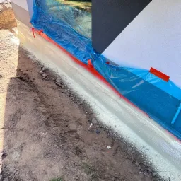 Świeżo nałożona, biała pianka izolacyjna na fundamencie budynku, zabezpieczona folią i czerwoną taśmą malarską, widoczna wykopana ziemia.