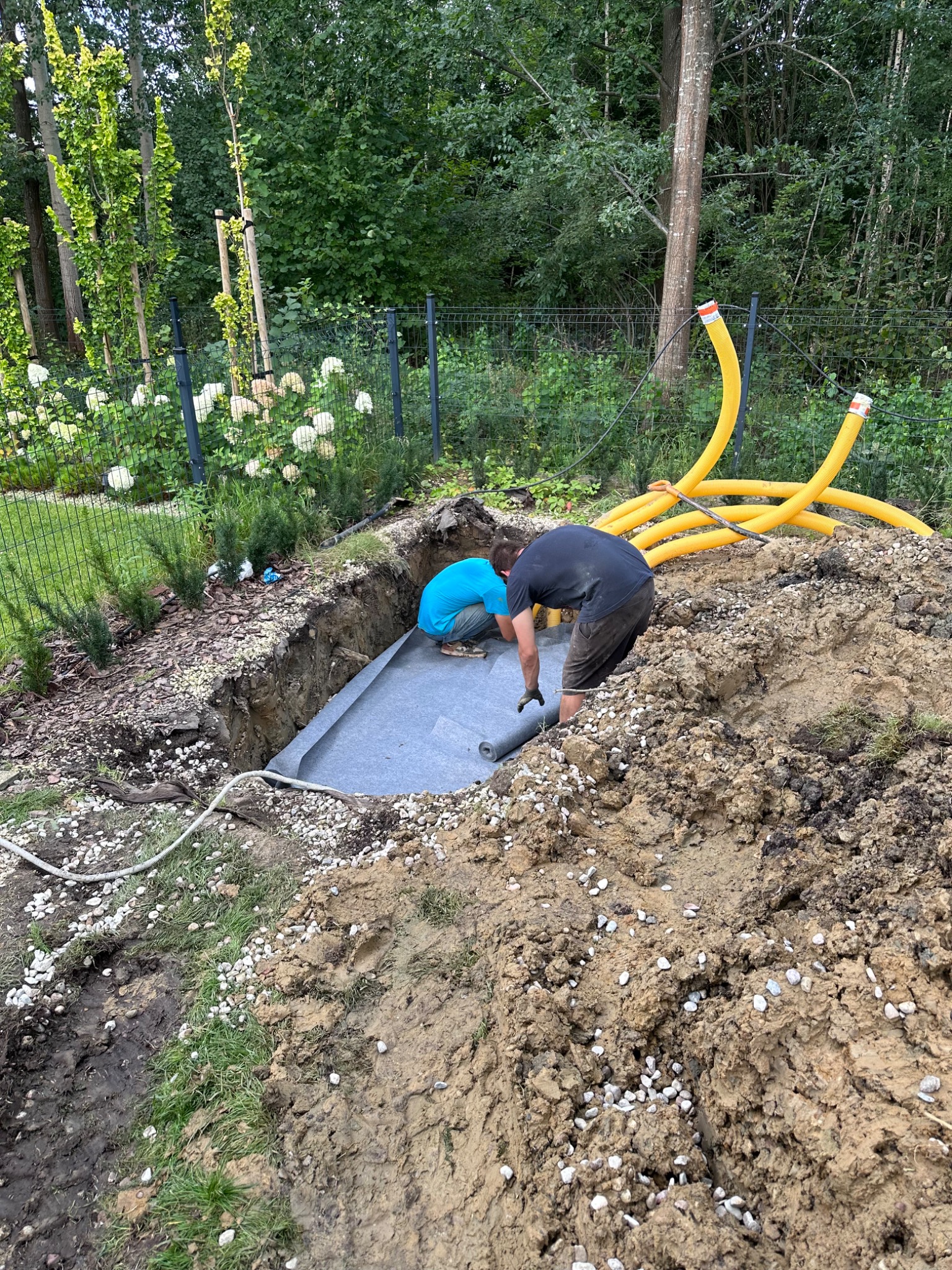 Dwóch mężczyzn rozkłada szarą membranę w wykopie pod fundamenty, widoczne żółte rury drenażowe, ziemia z kamieniami.