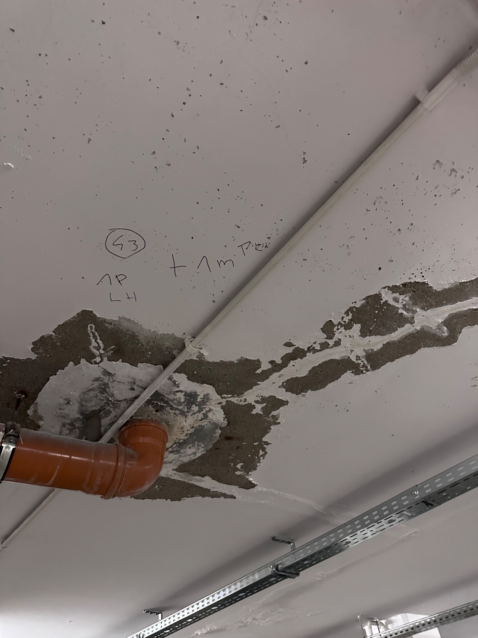 Uszkodzony sufit z widocznymi śladami wilgoci, naprawiany fragment z rurą kanalizacyjną i odręcznymi notatkami na suficie.