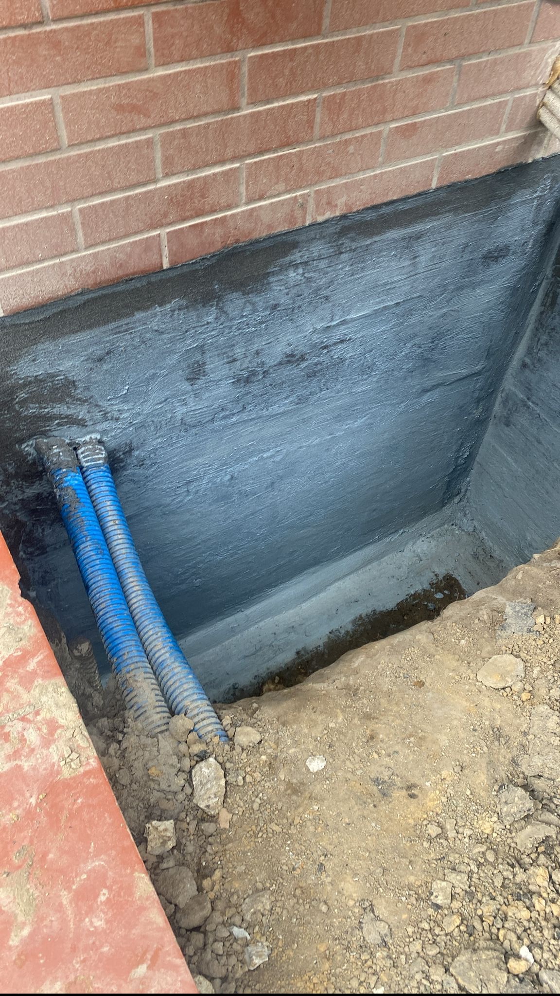 Świeżo zaizolowany fundament z widocznymi niebieskimi rurami karbowanymi ułożonymi w wykopie przy ceglanej ścianie.