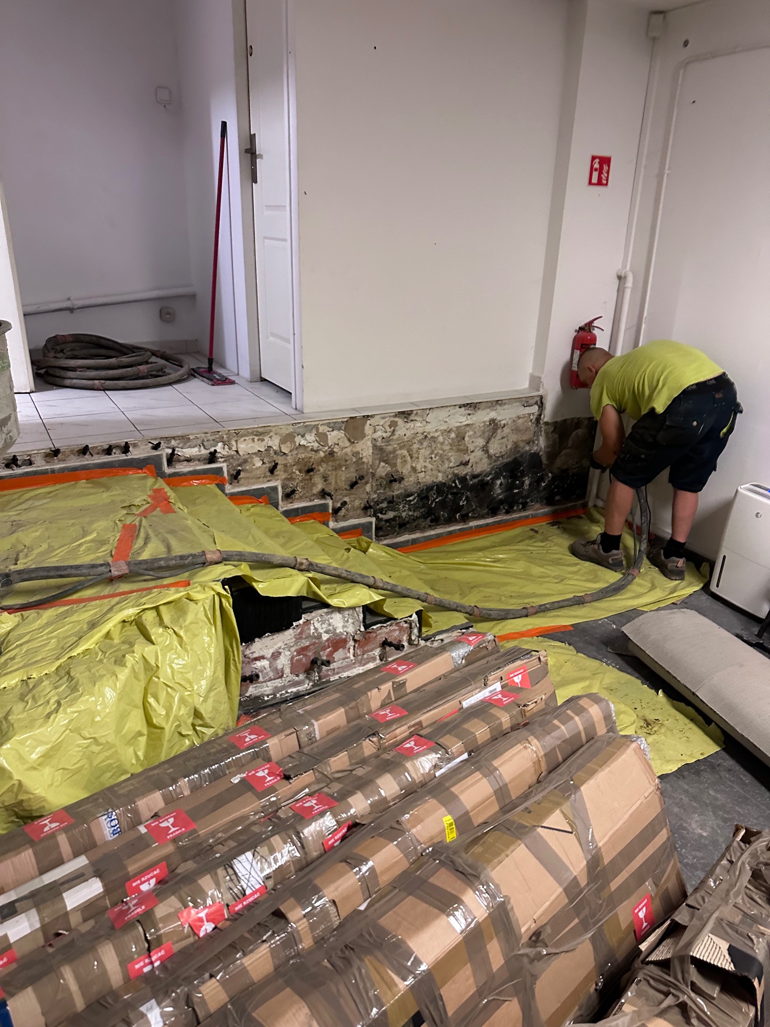 Prace przygotowawcze do osuszania zawilgoconej ściany w Trąbkach Wielkich, widoczne materiały budowlane i narzędzia na żółtej folii ochronnej, pracownik w trakcie działania przy ścianie.