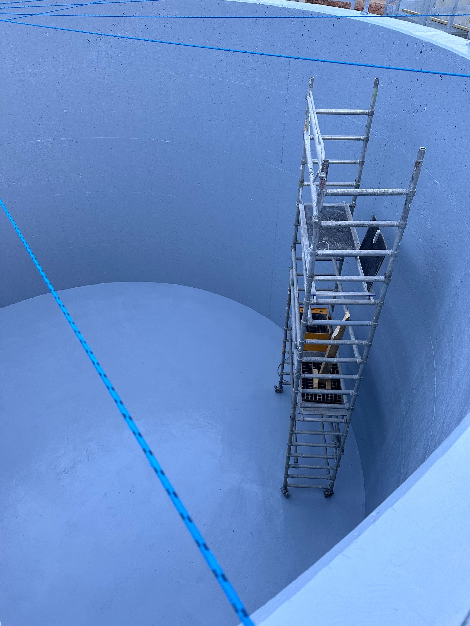 Wnętrze świeżo wyizolowanego, okrągłego zbiornika z metalową drabiną na kółkach, zabezpieczone niebieską liną.