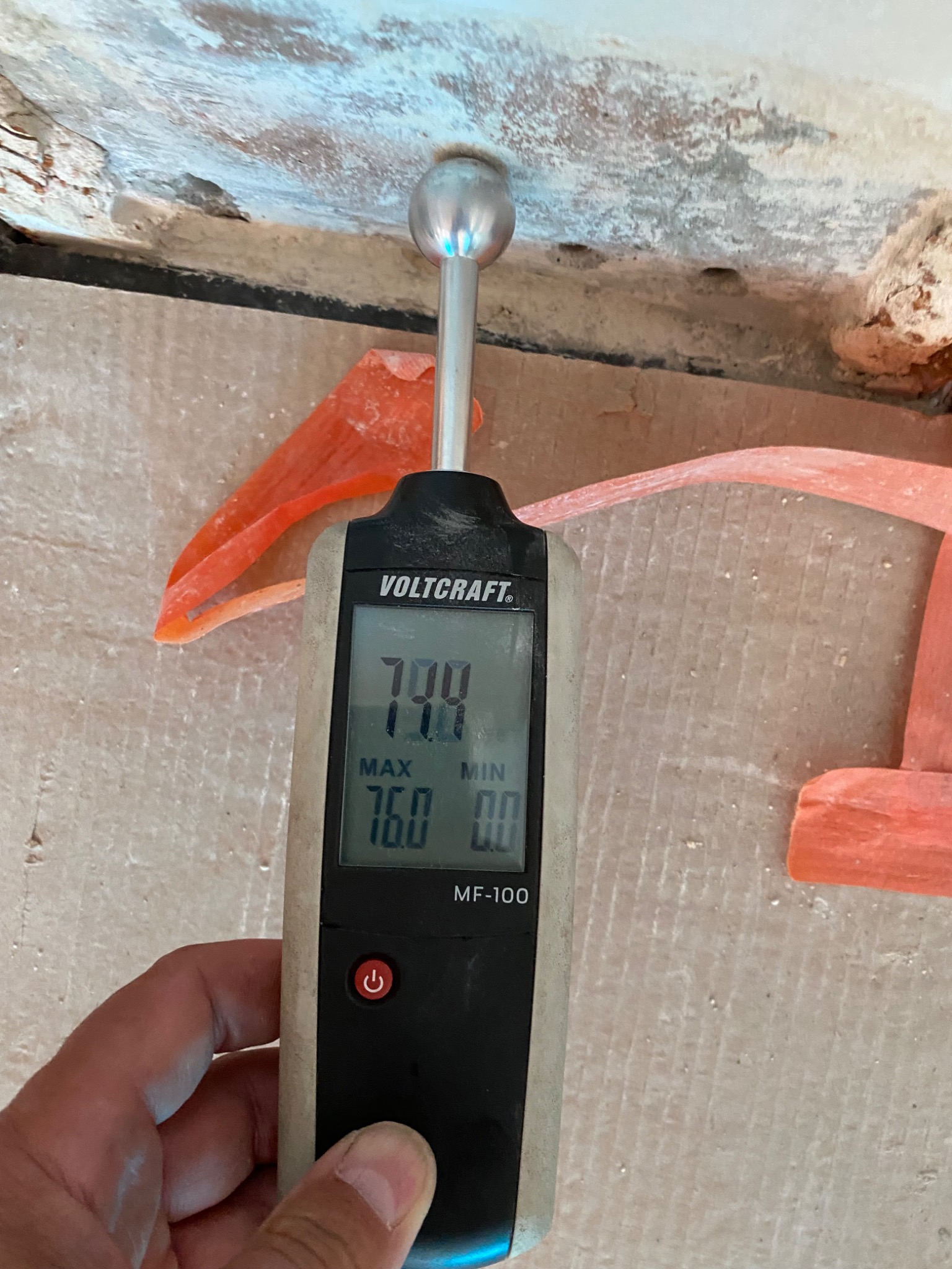 Miernik wilgotności Voltcraft MF-100, wskazujący 79.4%, przyłożony do ściany z widocznymi śladami zawilgocenia, z fragmentami pomarańczowej folii w tle.