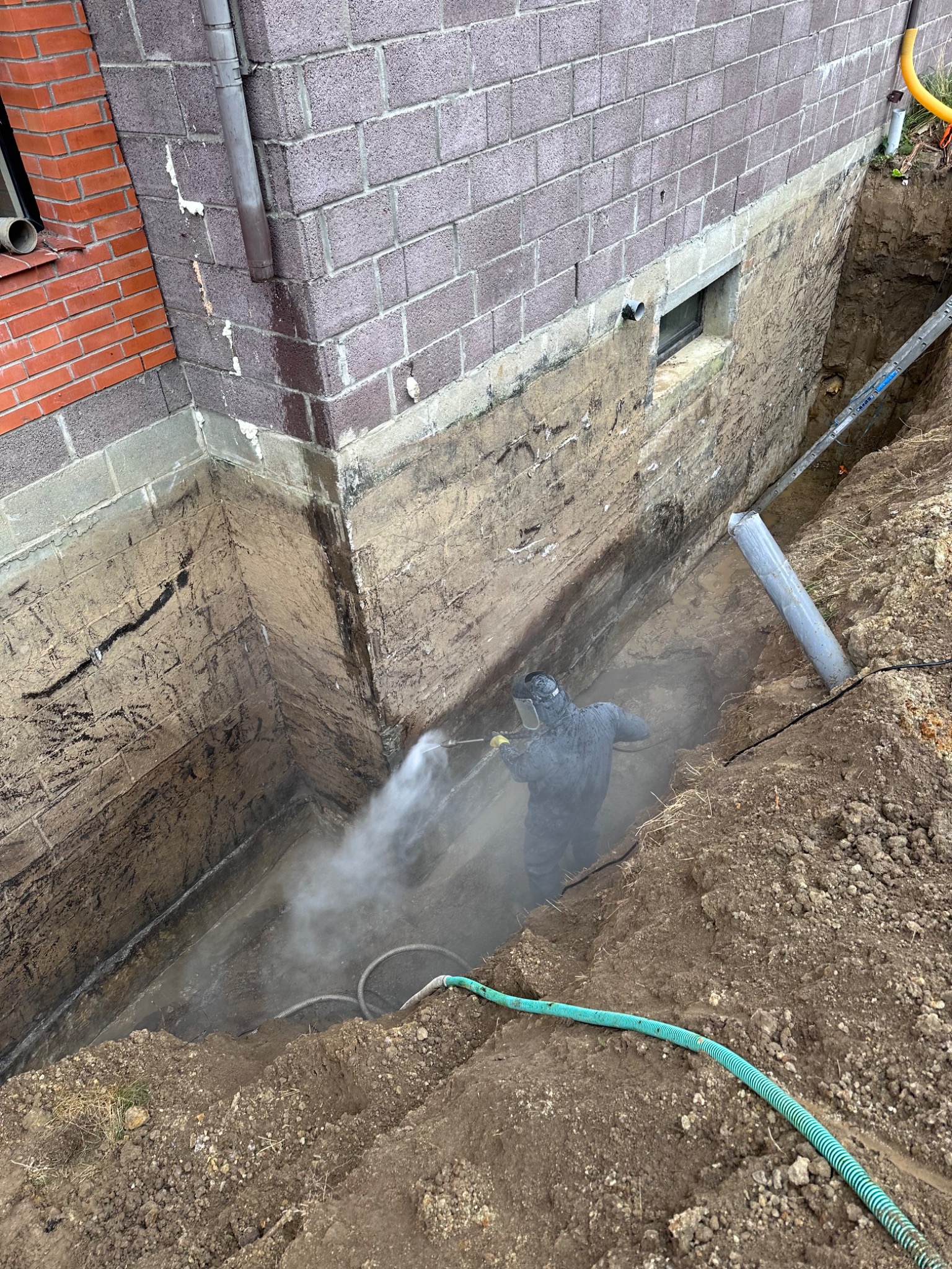 Pracownik w kombinezonie ochronnym czyści ścianę fundamentu budynku z cegły silikatowej po wykopaniu, przygotowując ją do izolacji; widoczny wąż i dysza pod ciśnieniem.