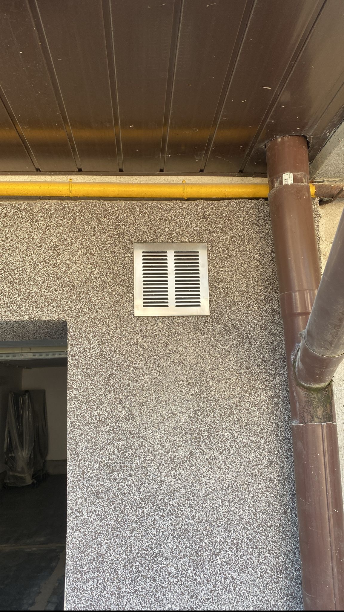 Ściana budynku pokryta tynkiem mozaikowym z kratką wentylacyjną i rurą spustową, widoczna żółta rura gazowa pod zadaszeniem.